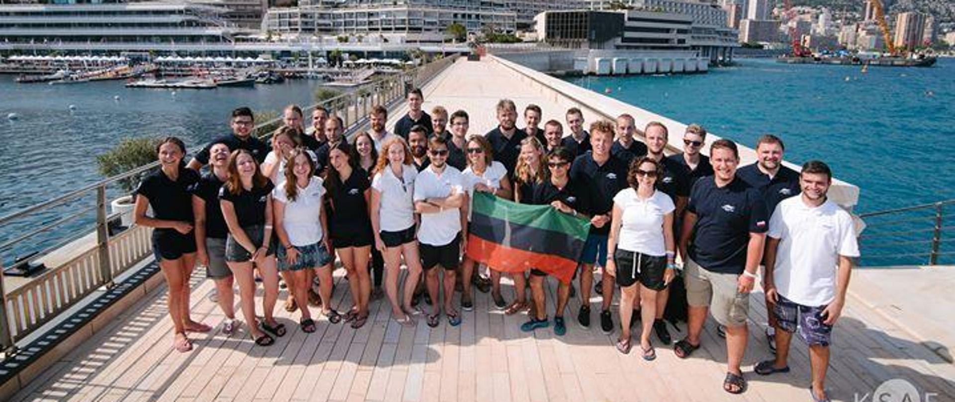 Grupa studentów stoi na tle długiego pomostu, w tle wybrzeże morza