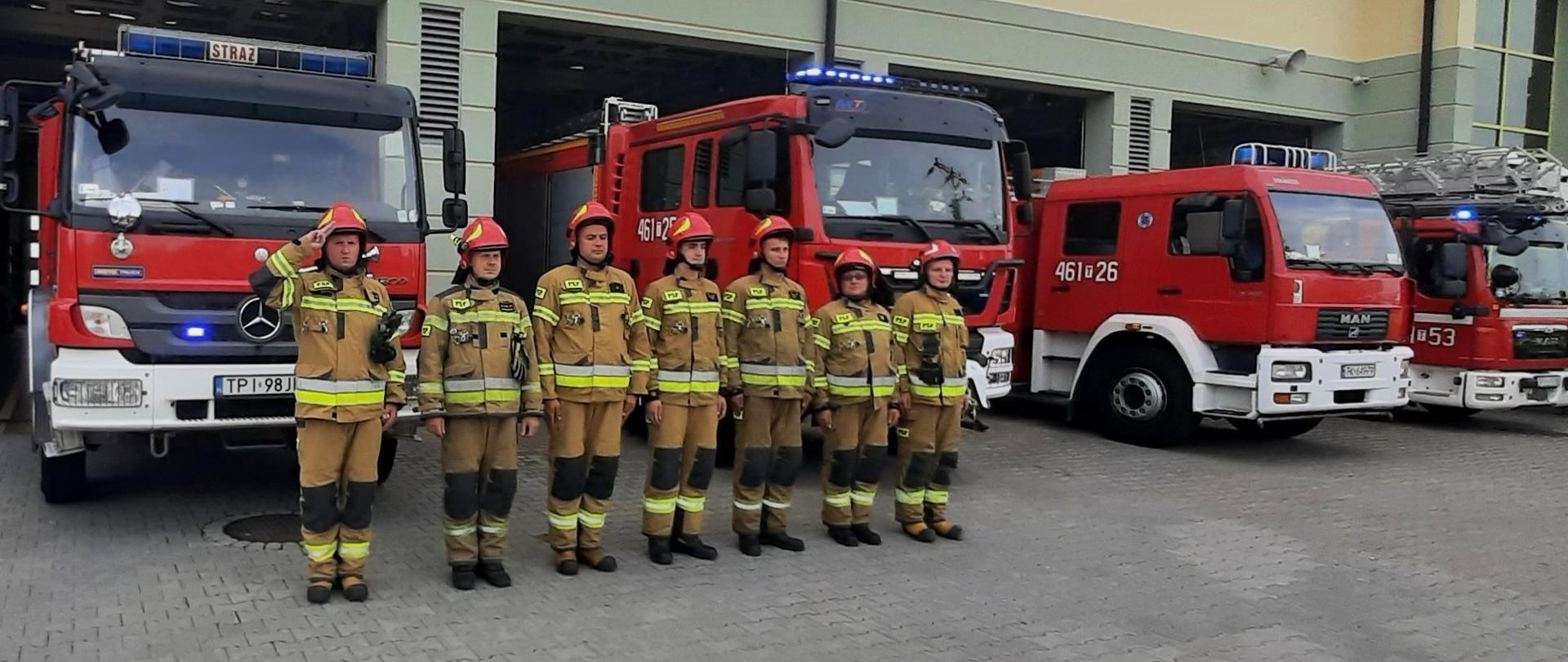 Strażacy w umundurowaniu specjalnym oddają hołd bohaterom Powstania Warszawskiego. Strażacy stoją w postawie zasadniczej na tle pojazdów pożarniczych. 