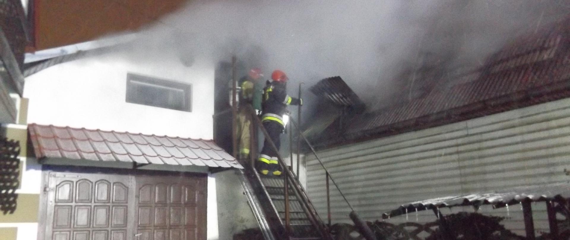 Zdjęcie przedstawia strażaków znajdujących się na schodach prowadzących na poddasze budynku mieszkalnego. Wokół panuje zadymienie. Strażacy prowadzą rozpoznanie.