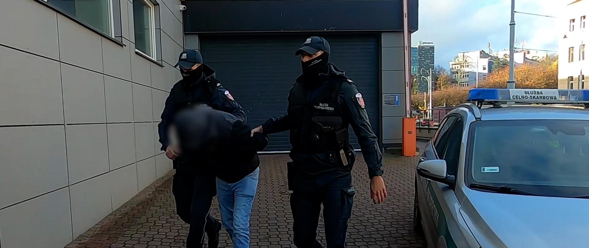 Funkcjonariusze prowadzą zatrzymanego, obok radiowóz Służby Celno-Skarbowej.