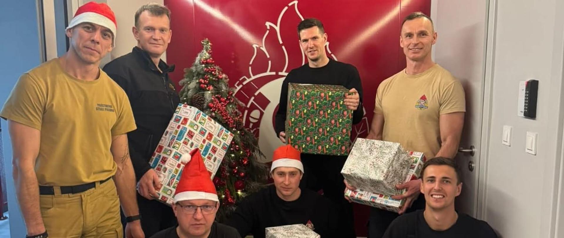 Na zdjęciu Zmiana Pierwsza z Jednostki Ratowniczo Gaśniczej pozuje do wspólnego zdjęcia. Strażacy trzymają w rękach spakowane prezenty, ubrani są w ubrania służbowe. 