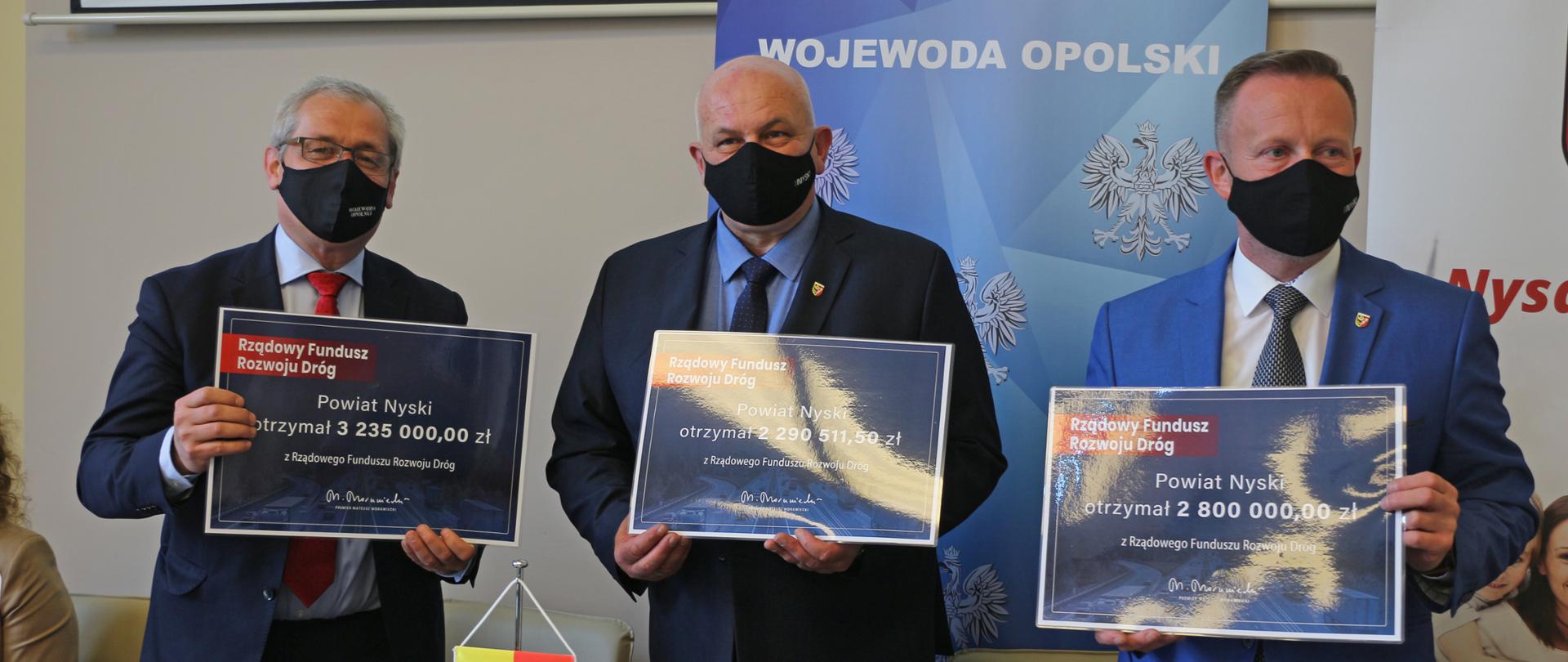 Na zdjęciu od lewej: wojewoda opolski oraz starosta nyski i wicestarosta nyski. Obaj pozują do zdjęcia. Stoją blisko siebie, w dłoniach trzymają trzy granatowe, symboliczne czeki. Ubrani w ciemne marynarki. W tle roll up gminy i wojewody. Przed nimi stół, na którym stoją wody mineralne i flagietki z herbem Nysy. 