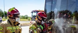 Dwóch strażaków Państwowej Straży Pożarnej prowadzi działania ratownicze przy autobusie. Strażak znajdujący się na pierwszym planie, wyposażony w hełm, aparat ochrony dróg oddechowych i ubranie specjalne, przy użyciu akumulatorowego narzędzia ratowniczego wykonuje dostęp przez szybę pojazdu. Drugi ratownik zabezpiecza prowadzone działania. Na szybie autobusu widoczne są uszkodzenia powstałe w wyniku prowadzonej pozoracji. W tle znajdują się samochody ratownicze oraz pozostali uczestnicy ćwiczeń.