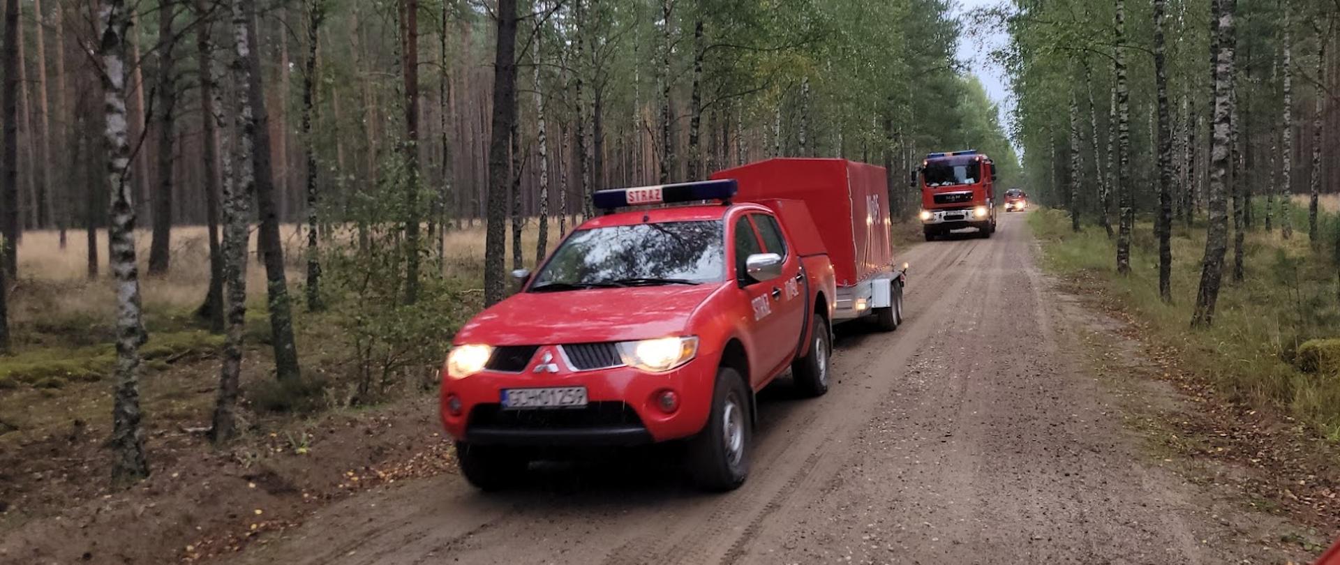 Doga leśna, drogą poruszają się samochody strażackie. Na pierwszym planie samochód typu pickup ciągnie przyczepkę sprzętową. W tle większy samochód strażacki. 