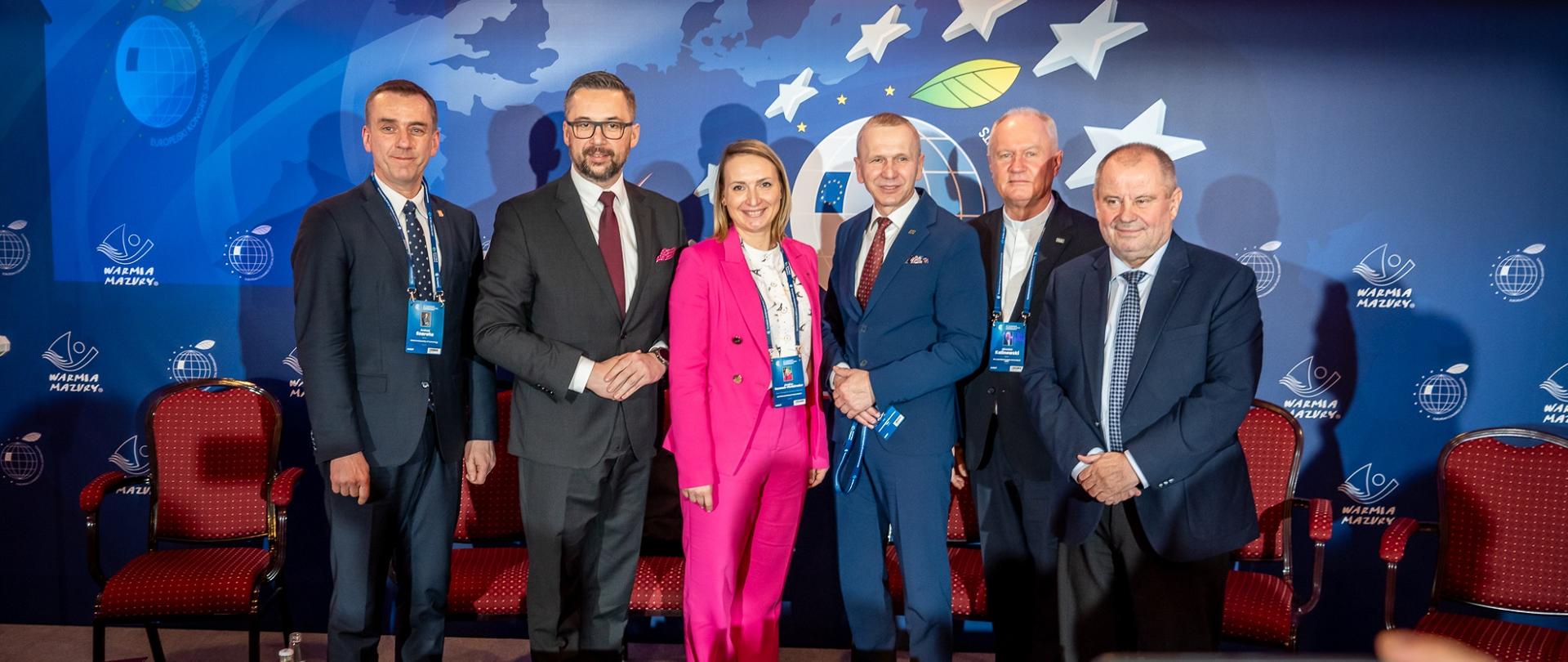 Na tle niebieskiej ściany z napisem Warmia i Mazury stoi w szeregu sześć osób, pięciu mężczyzn w garniturach i kobieta w różowym ubraniu.