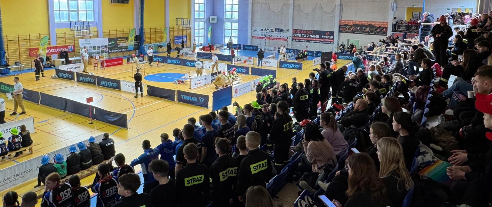 Strażacy podczas wykonywania ćwiczeń na torze. Na trybunach siedzą widzowie, strażacy czekający na swój start. Strażacy podczas dekoracji, z włodarzami i przedstawicielami instytucji.