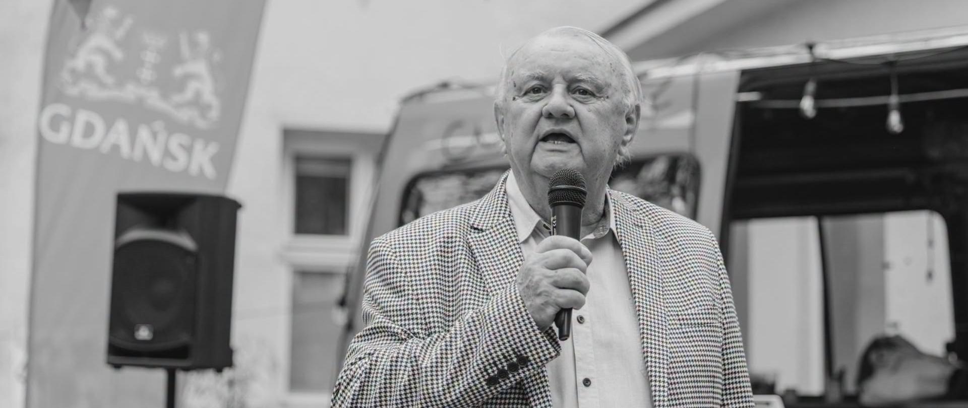 Na zdjęciu widać mężczyznę branego w jasną koszulę oraz wzorzystą marynarkę, który trzyma w dłoni mikrofon. Cała fotografia zachowana jest w czarno-białej tonacji. W tle można zauważyć budynek baner z napisem Gdańsk oraz herbem miasta, a także głośnik na statywie.

