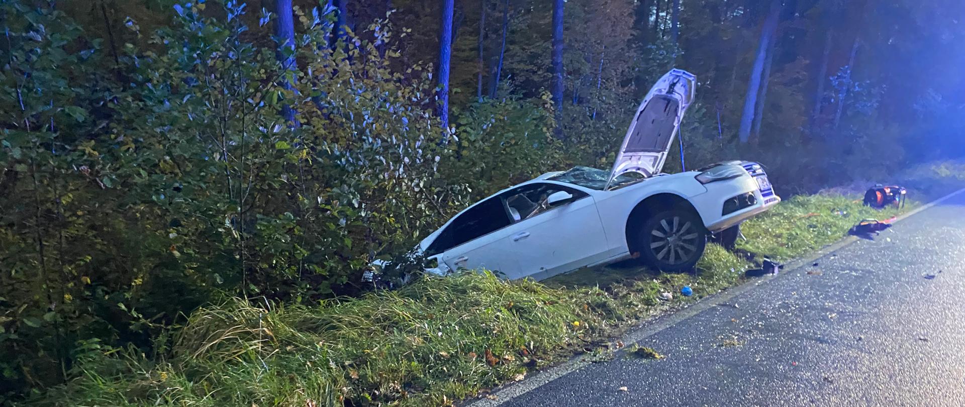 Na poboczu drogi widać biały samochód osobowy, który wpadł do rowu. Przód pojazdu znajduje się w zaroślach, a tylna cześć uniesiona jest do góry. Maska samochodu jest otwarta. Na drodze widoczne są fragmenty karoserii i ślady po kolizji.