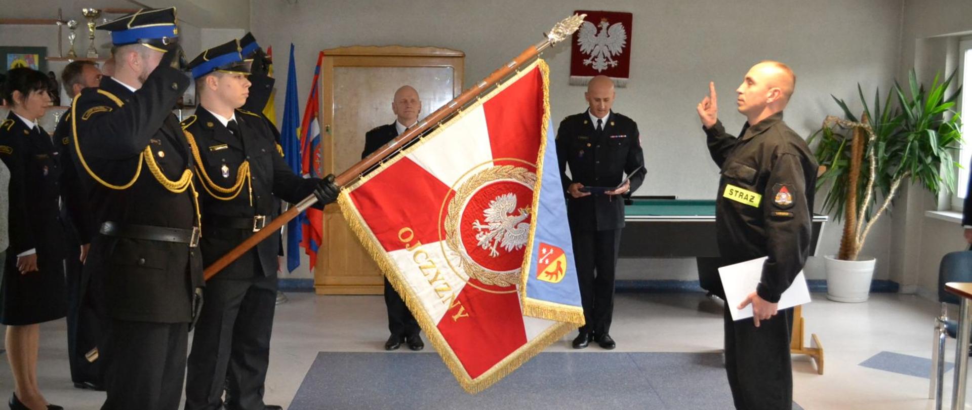 Na pierwszym planie widoczny jest strażak składający ślubowanie przy poczcie sztandarowym. Poczet składa się z trzech strażaków z czego środkowy trzyma opuszczony poczet. W tle widoczne są osoby uczestniczące w apelu.