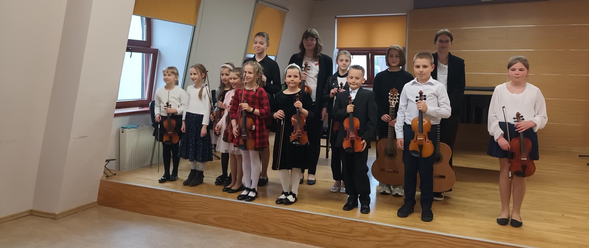 Uczestnicy audycji sekcji strunowej stoją w dwóch rzędach na scenie sali kameralnej, trzymając w dłoniach w pozycji pionowej swoje instrumenty. Z lewej strony dwa okna przysłonięte roletami, za nimi dwa fortepiany oraz jedno okno przysłonięte roletą.