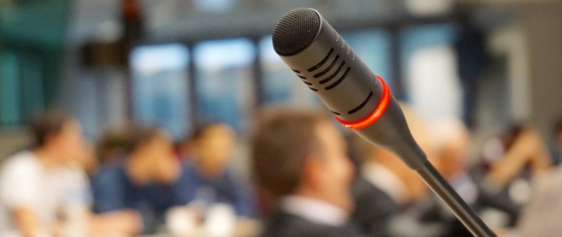 Włączony mikrofon z czerwoną obwódką świetlną, w tle widoczni rozmyci uczestnicy spotkania.