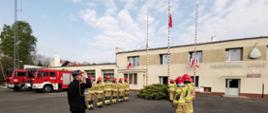 Strażacy PSP w ubraniach specjalnych oraz w mundurze wyjściowym na placu JRG Paczków podczas uroczystej zbiórki i podniesienia Flagi Rzeczypospolitej Polskiej z okazji święta 2 maja. W tle widoczne samochody ratowniczo-gaśnicze przed garażami jednostki.