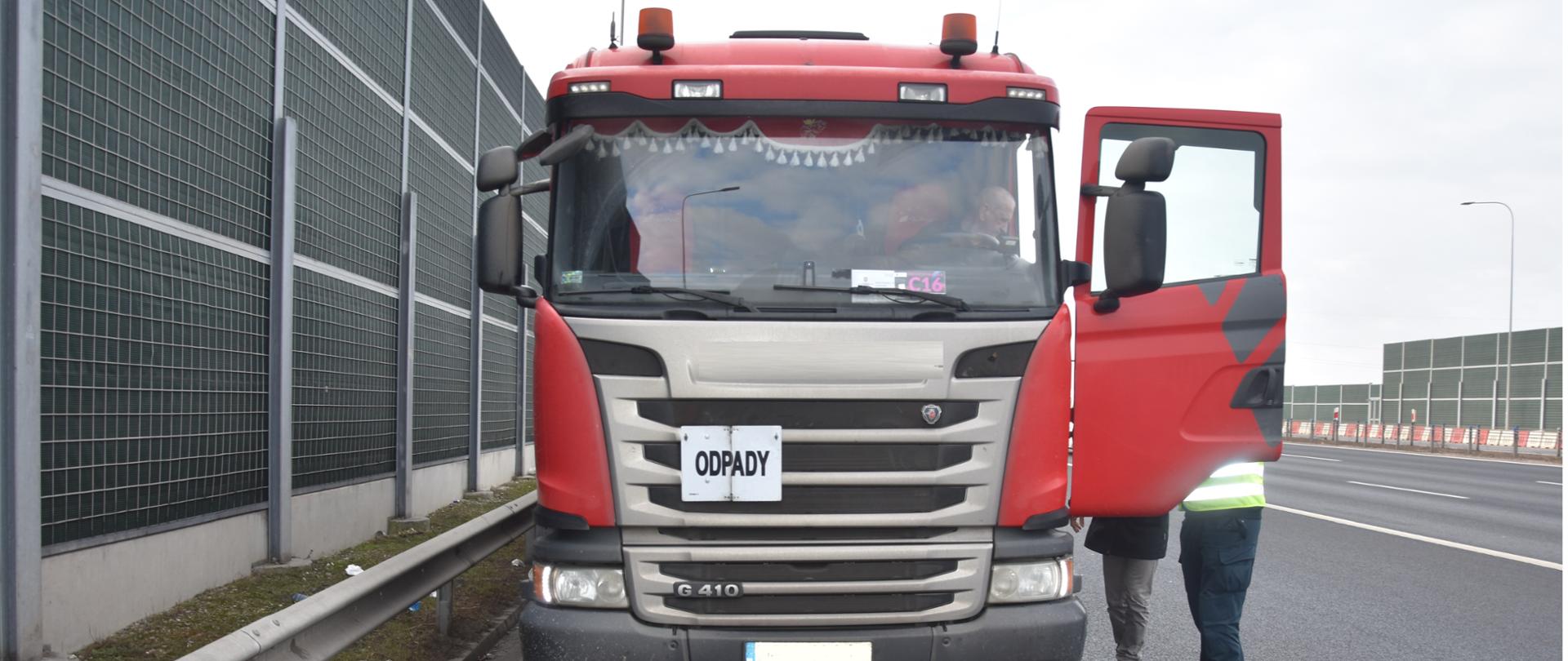 Inspektor Wojewódzkiego Inspektoratu Ochrony Środowiska w Warszawie kontroluje samochód ciężarowy stojący na poboczu wielopasmowej drogi szybkiego ruchu. Po lewej stronie widać zielony ekran akustyczny.