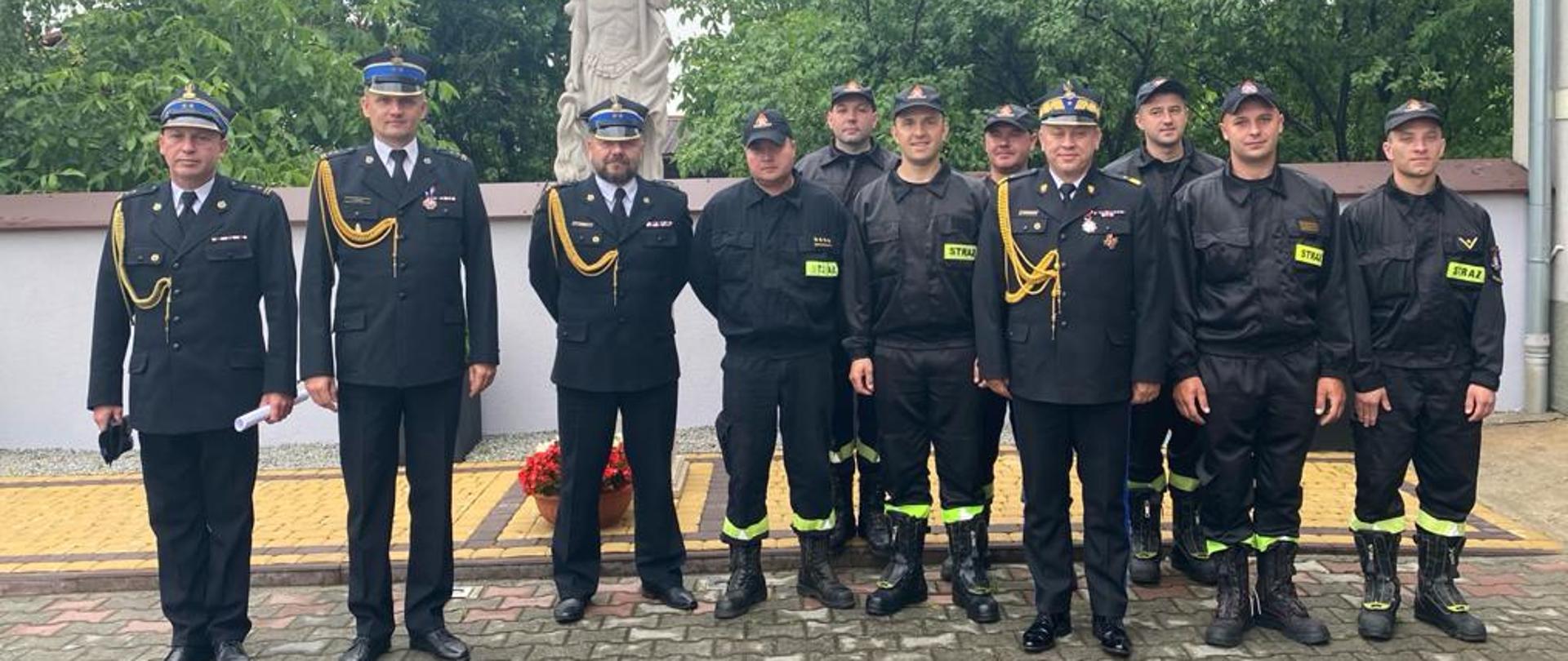 Fotografia przedstawiająca Mazowieckiego Komendanta Wojewódzkiego wraz Komendantem Powiatowym PSP oraz załogą JRG Szydłowiec stojących na tle św. Floriana 