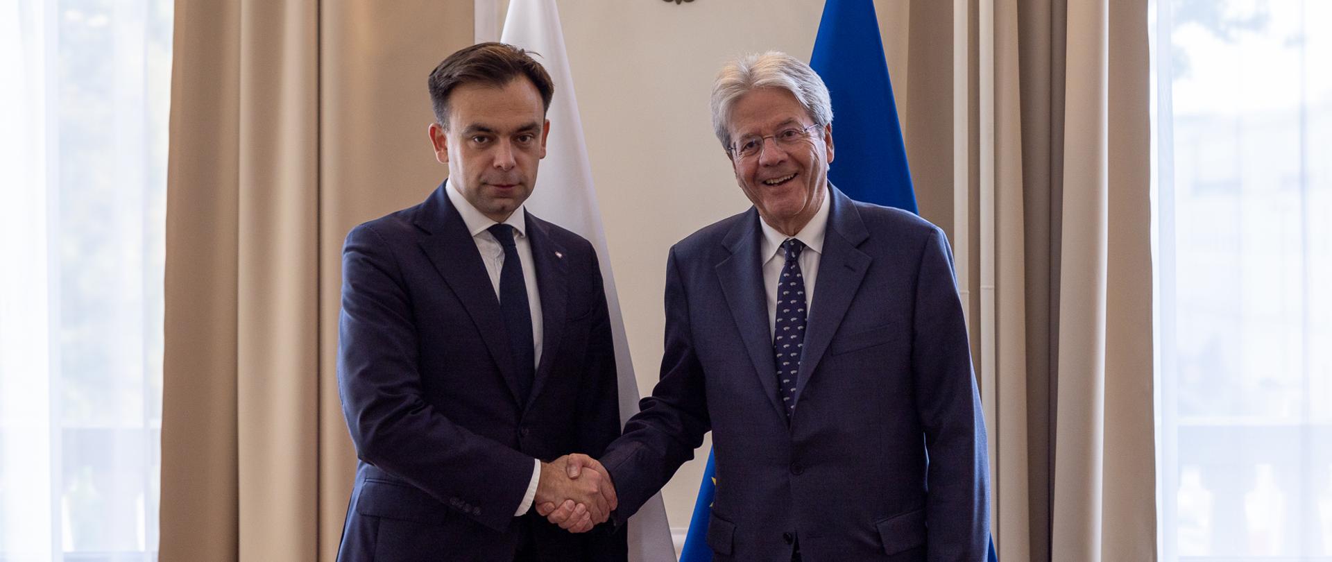 Minister finansów Andrzej Domański podaje rękę Komisarzowi UE ds. gospodarki Paolo Gentilonim, w tle flagi Polski i flaga Unii Europejskiej