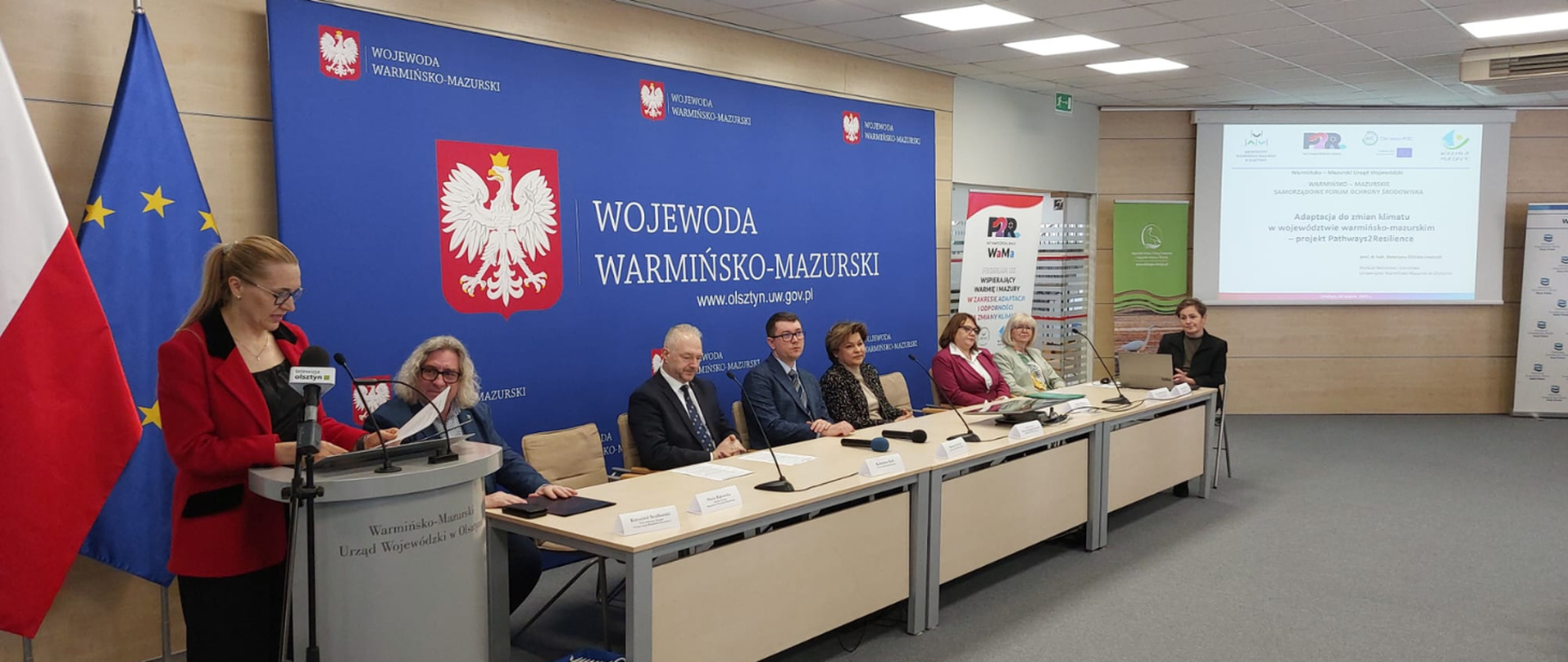 Duży stół, na którym stoją mikrofony oraz leżą dokumenty. Przy stole siedzą ludzie i słuchają wystąpienia pani stojącej przy mównicy.