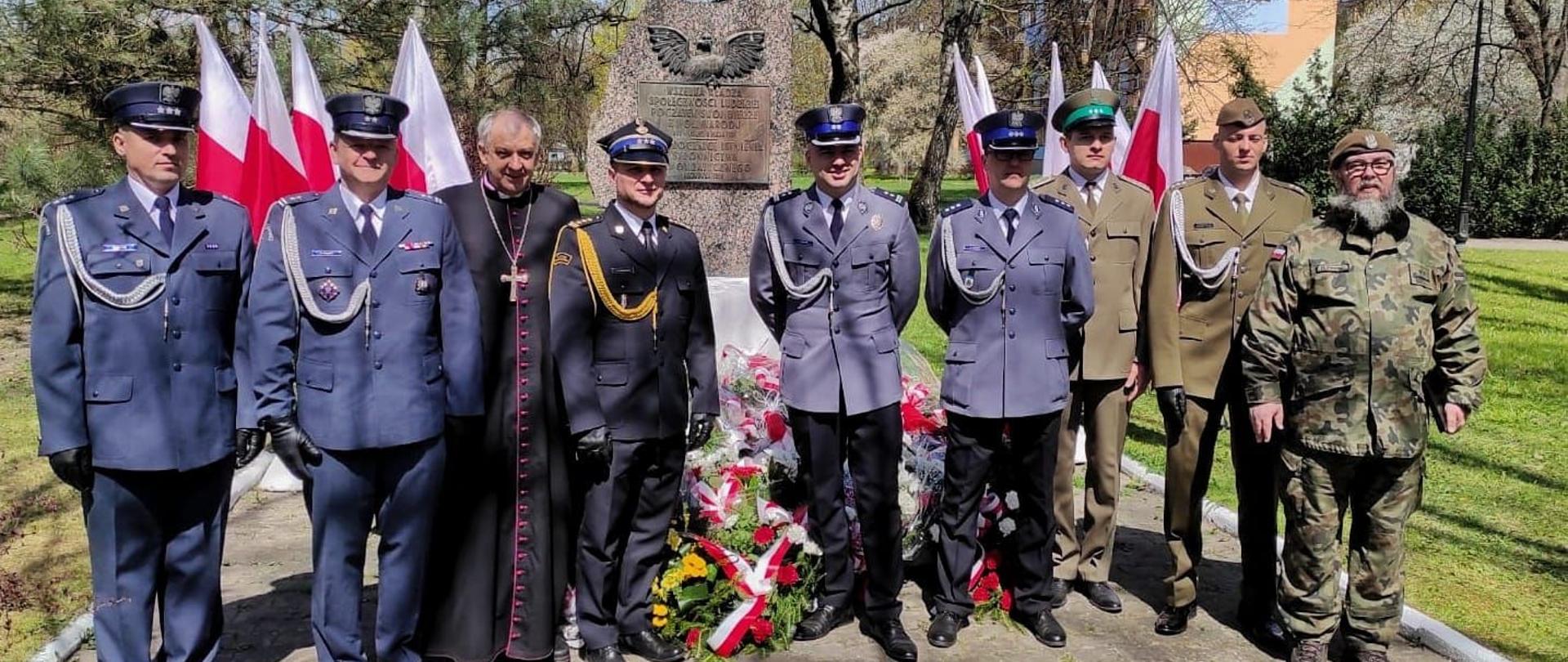 Przed pomnikiem w rzędzie stoi 10 osób. Ksiądz w sutannie oraz dziewięciu funkcjonariuszy w różnych mundurach. za nimi wiązanki kwiatów.