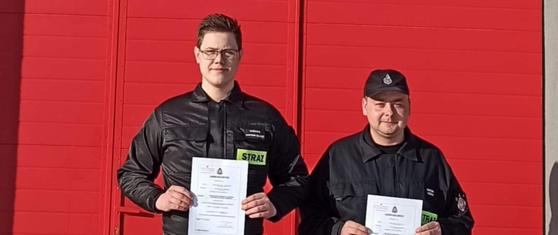 Na tle czerwonej bramy garażowej remizy OSP stoją dwaj druhowie z OSP ubrani w ubrania koszarowe. W dłoniach trzymają zaświadczenie formatu A4 potwierdzające ukończenie szkolenia podstawowego.
