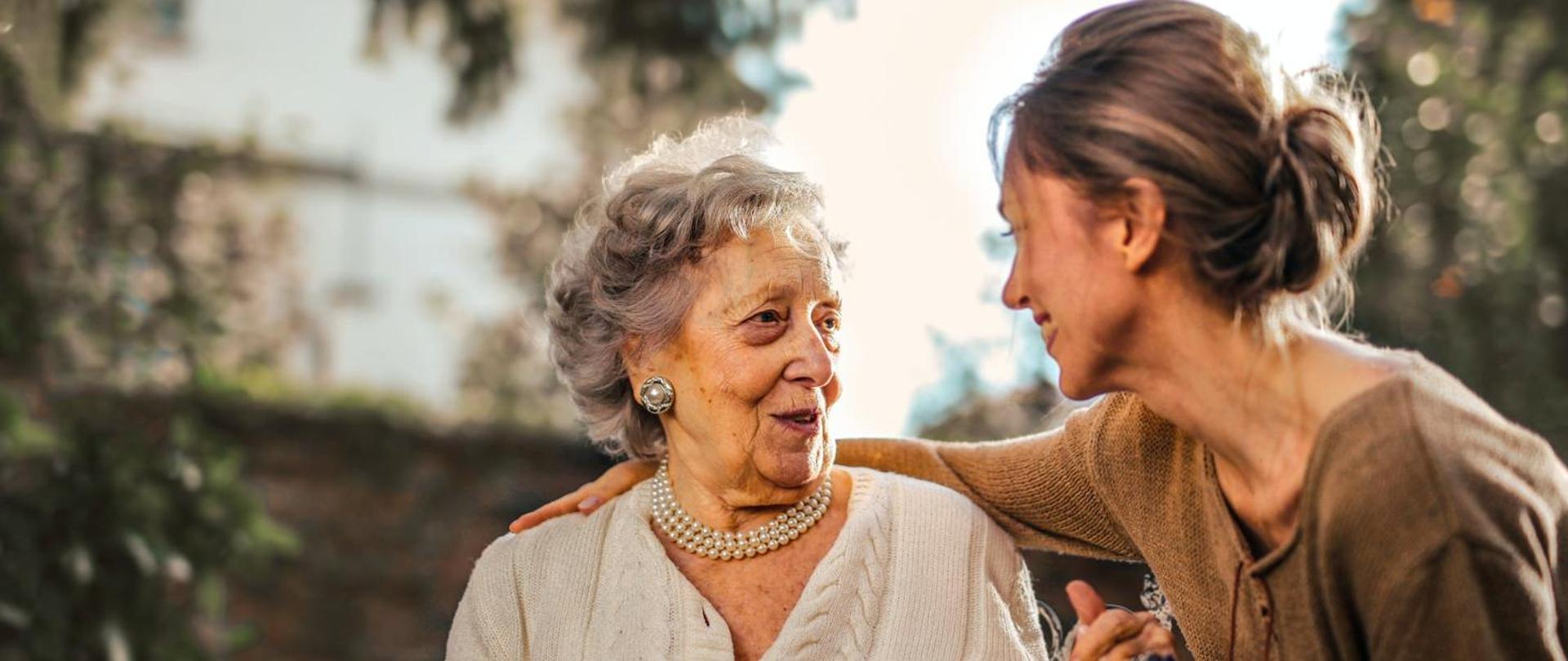 Na zdjęciu starsza kobieta ubrana w jasny sweter. Ma na sobie kolczyki oraz perły na szyi. Uśmiecha się do młodej kobiety, która położyła rękę na jej plecach. Młoda kobieta ubrana jest w brązowy sweter. Widać ją profilem. 