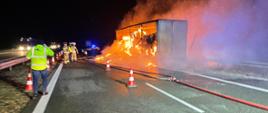 Naczepa samochodowa stoi na poboczu drogi w środku kostki papieru objęte ogniem - cała naczepa w płomieniach, wzdłuż naczepy linia wężowa oraz ułożone pachołki na pierwszym planie pan w żółtej kamizelce rozmawia przez telefon na drugim planie strażacy oraz policjant stoją przy białej linii odznaczająca pas autostrady na drugim pasie widać światła przejeżdżających samochodów