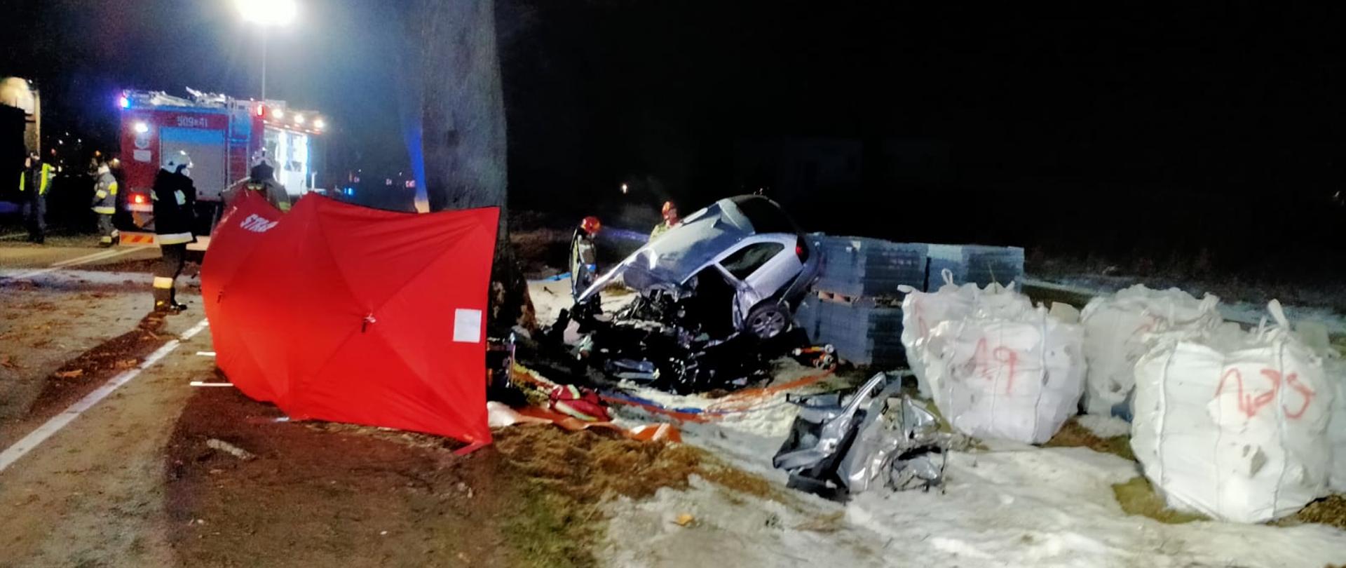 Samochód koloru srebrnego poza jednią z widocznym rozbitym przodem po uderzeniu w drzewo, bok rozłożony czerwony parawan zakrywający część pojazdu. W tle strażacy oraz samochód straży pożarnej z włączoną lampą oświetlający teren prowadzonej akcji.