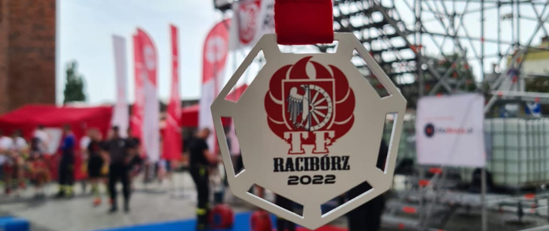 Na zdjęciu widać medal za udział w zawodach z napisem Racibórz 2022. W tle widać zamazany obraz ścianki na która podczas zawodów wchodzą zawodnicy. 