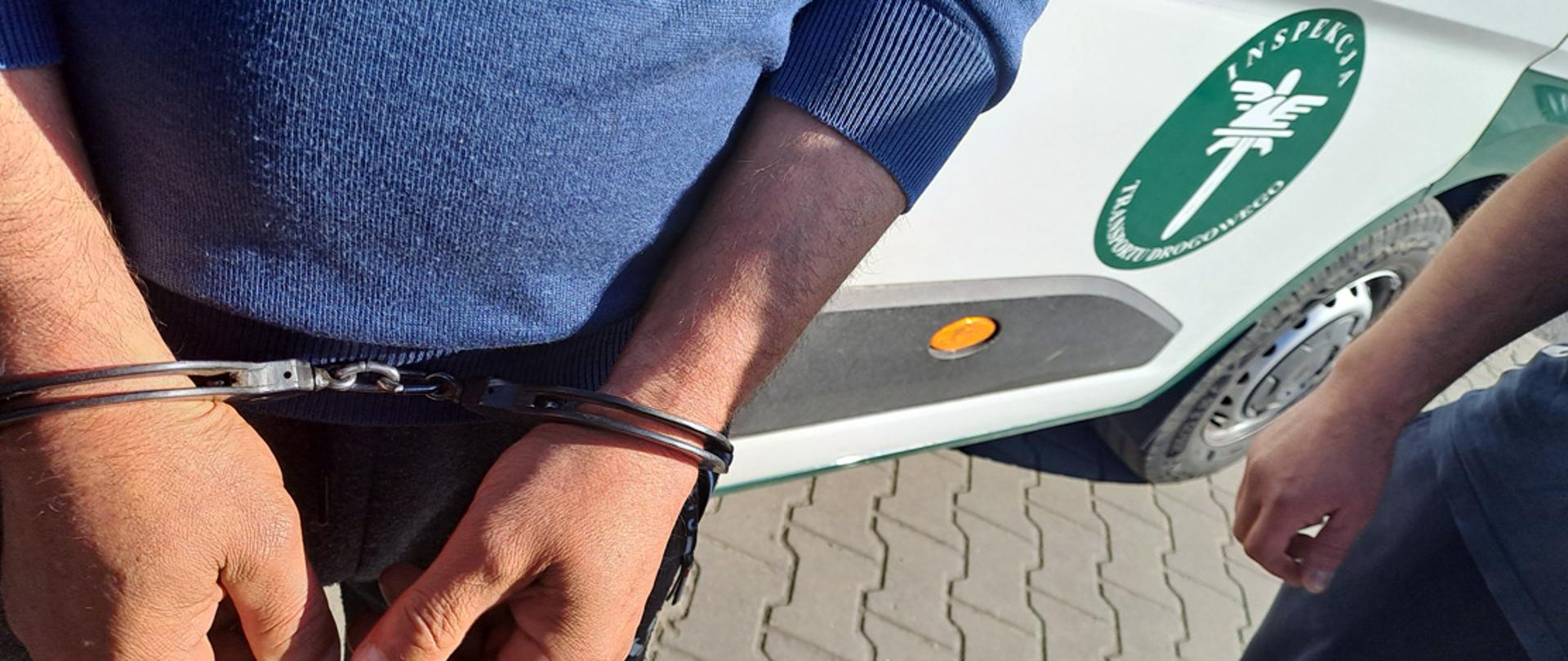 Mężczyzna z założonymi kajdankami na rękach stoi przed samochodem służbowym Inspekcji Transportu Drogowego