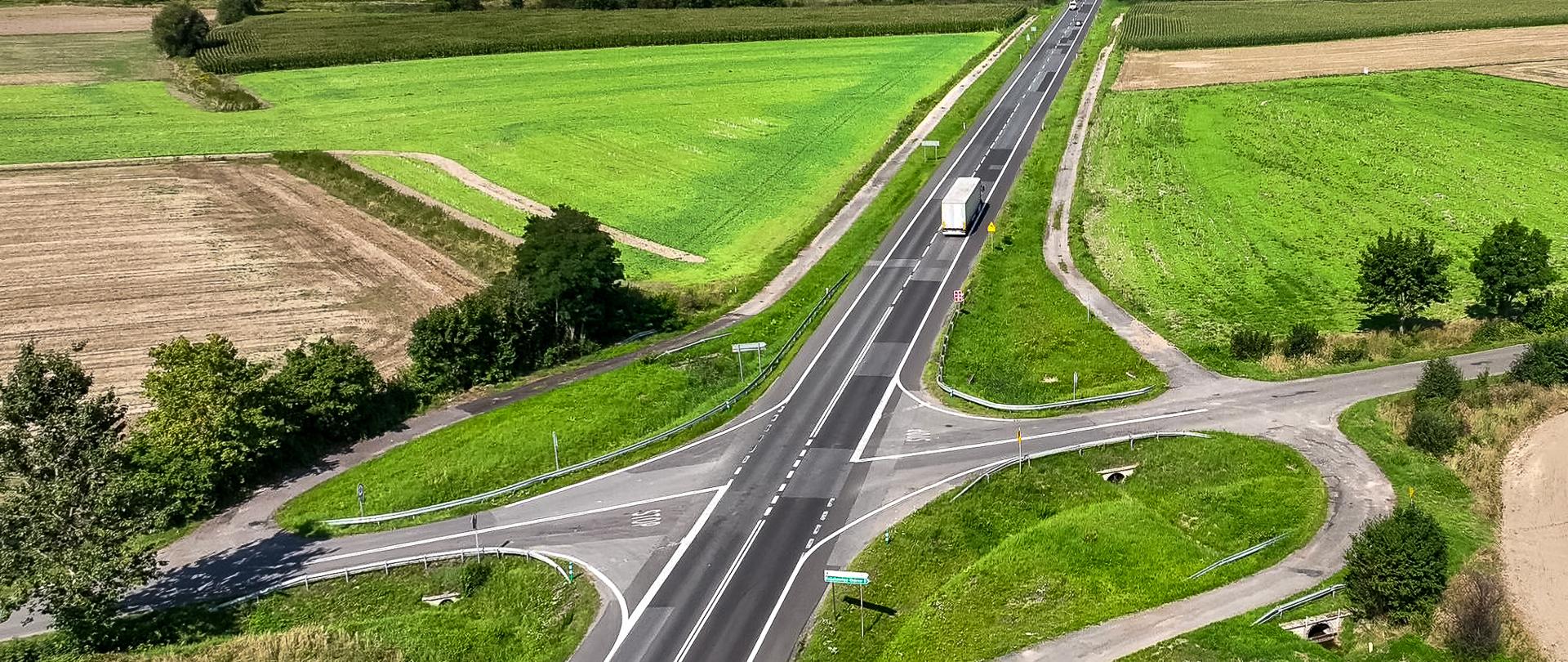 Dwupasowa jezdnia, umiarkowany ruch pojazdów. Po obu stronach drogi łąki i pola uprawne. W oddali widoczne zabudowania