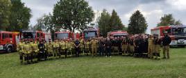 Zdjęcie przedstawia strażaków podczas ćwiczeń.
W tle samochody tereny zielone.
