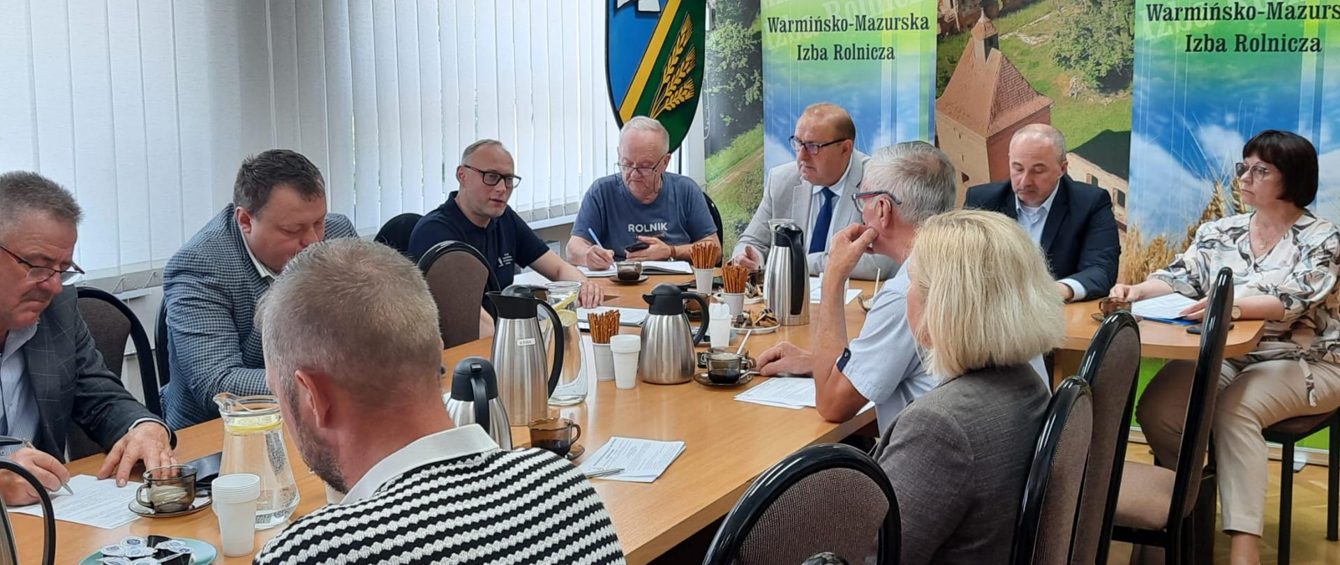 spotkanie uczestników Rady Powiatowej Izb Rolniczych przy długim stole konferencyjnym. W tle widoczne są banery Warmińsko-Mazurskiej Izby Rolniczej oraz herb Gminy Iława.
