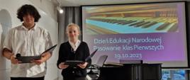 Yevhenii Oleksiienko i Wiktoria Habrajska w roli prowadzących. Uczniowie stoją na scenie. W rękach trzymają otwarte teczki. W tle widać fortepian i wyświetlony na ekranie slaid z grafiką klawiatury fortepianu i napisem Dzień Edukacji Narodowej oraz Pasowanie klas Pierwszych 19.10.2023
