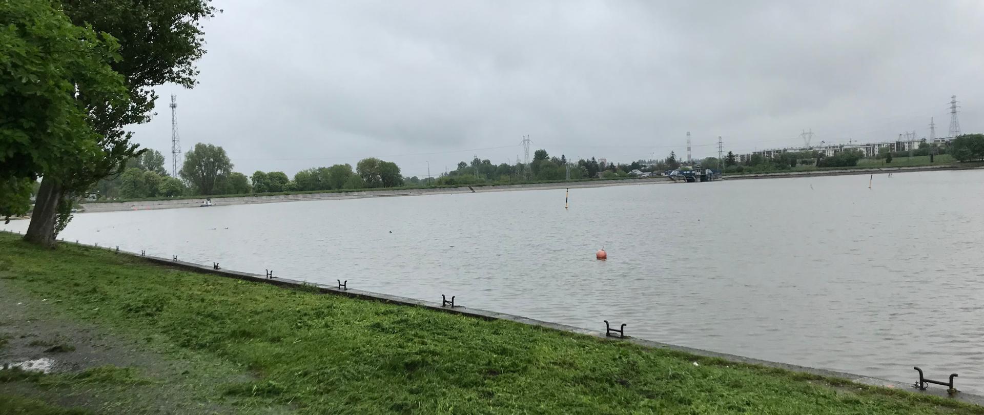 Zbiornik wodny. Na brzegu zielona trawa. Nad zbiornikiem zachmurzone niebo.