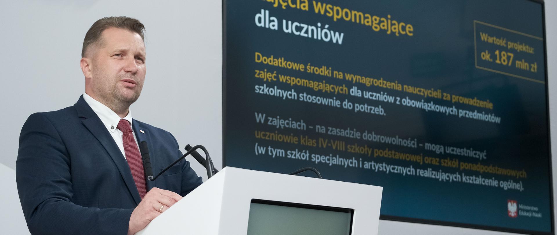 Minister Edukacji i Nauki Przemysław Czarnek stoi przy mównicy podczas konferencji. W tle ekran z prezentacją.