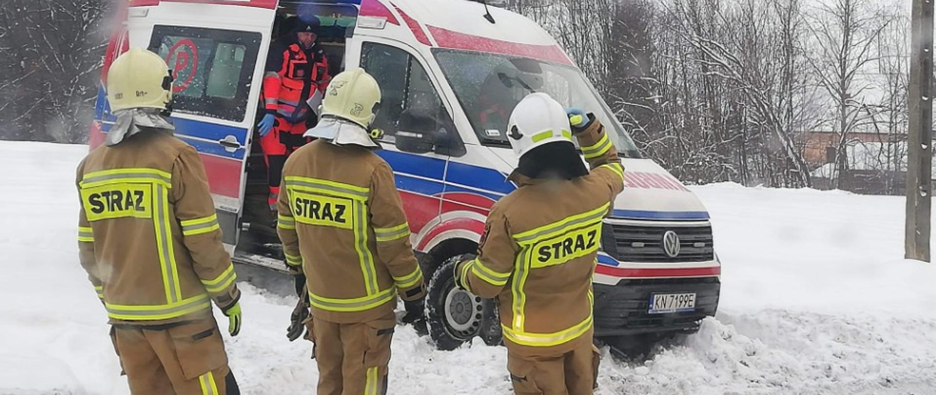 Trzech strażaków stoi obok karetki Pogotowia ratunkowego. Wszędzie leży dużo śniegu.