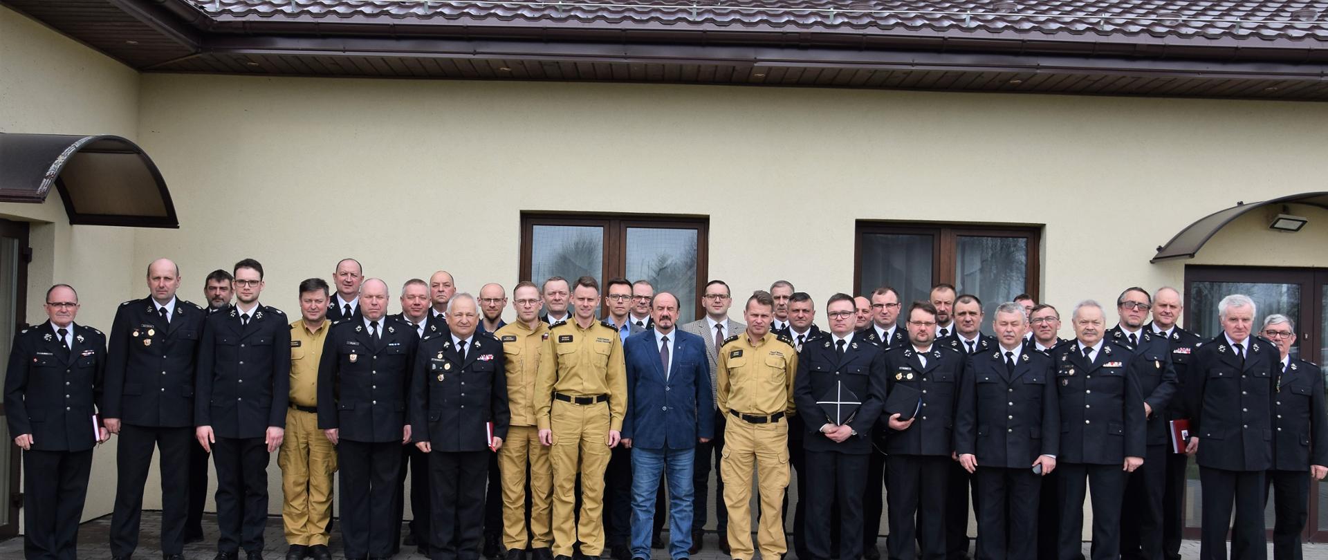 Wspólne zdjęcie uczestników spotkania. Strażacy OSP w mundurach wyjściowych, funkcjonariusz PSP ubrani w nowe umundurowania służbowe koloru piaskowego.