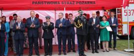Pod czerwonym ekspresowym namiotem zaproszeni goście. W pierwszym rzędzie od lewej: wójt gminy Marianowo Danuta Ankutowicz, poseł Michał Jach, poseł Leszek Dobrzyński, druh Stanisław Kopeć, Sekretarz Stanu Paweł Szefernaker, nadbrygadier Jarosław Tomczyk - komendant salutuje.