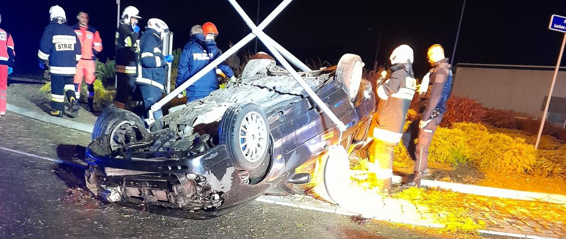 Samochód osobowy na rondzie leży na dachu. Obok strażacy.