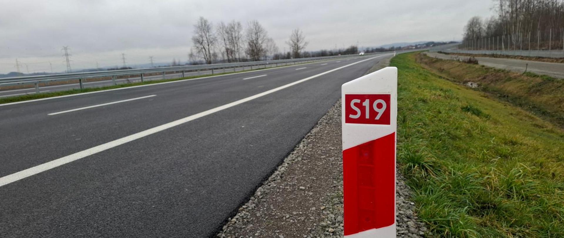 Zdjęcie pokazuje fragment dwupasmowej drogi ekspresowej S19 widziany z prawej strony jezdni. Na pierwszym planie stoi biało‑czerwony słupek kilometrowy z oznaczeniem „S19”. Po lewej stronie widać szeroki pas asfaltu i metalowe bariery, a po prawej zielone pobocze. W tle rozciągają się pola, pojedyncze drzewa i szare, pochmurne niebo.