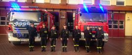 Strażacy stoją w ubraniach koszarowych przed wozami strażackimi, które mają włączone sygnały świetlne. W tle budynek straży pożarnej.