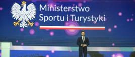 Minister Kamil Bortniczuk wziął udział w Noworocznym Spotkaniu Rodziny Olimpijskiej. Minister Bortniczuk stoi na scenie, mówi do mikrofonu, za nim na ścianie LED logotyp Ministerstwa Sportu i Turystyki