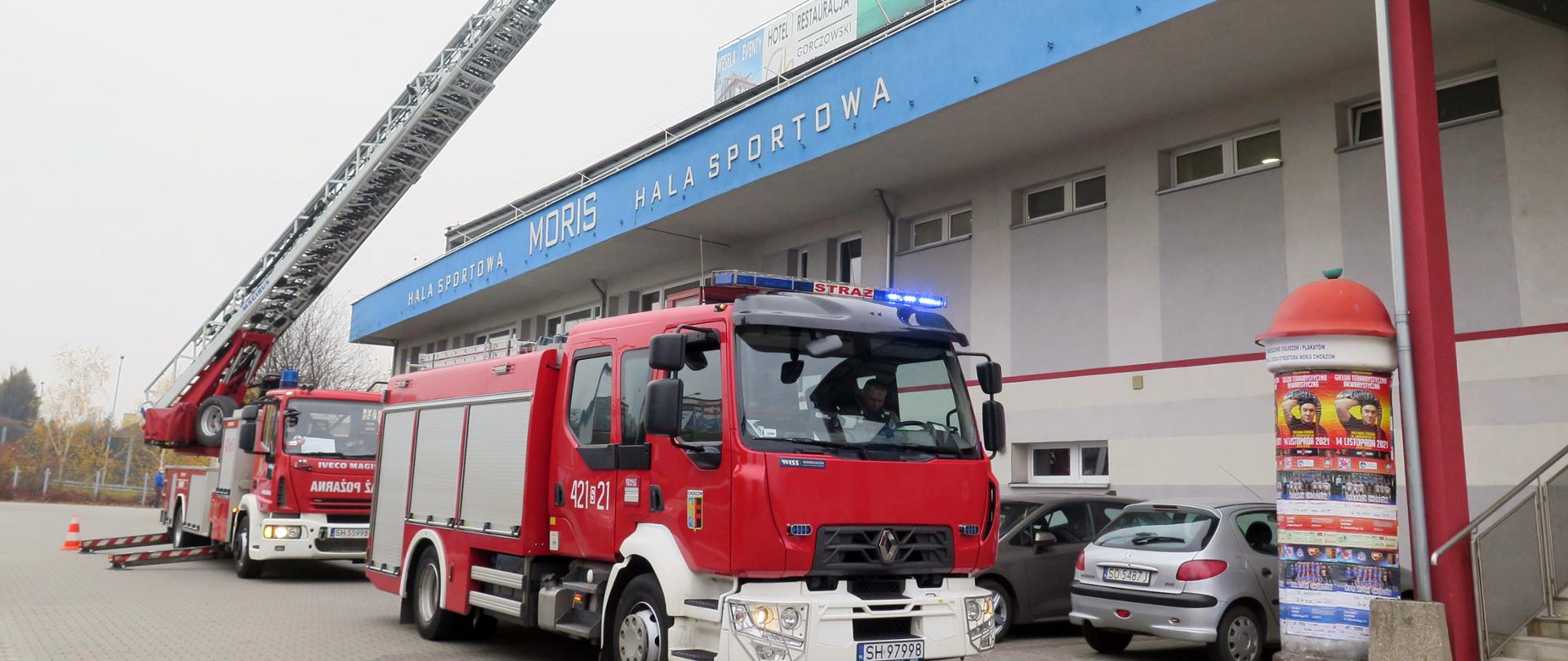Zdjęcie przedstawia dwa samochody pożarnicze stojące na tle hali sportowej MORiS