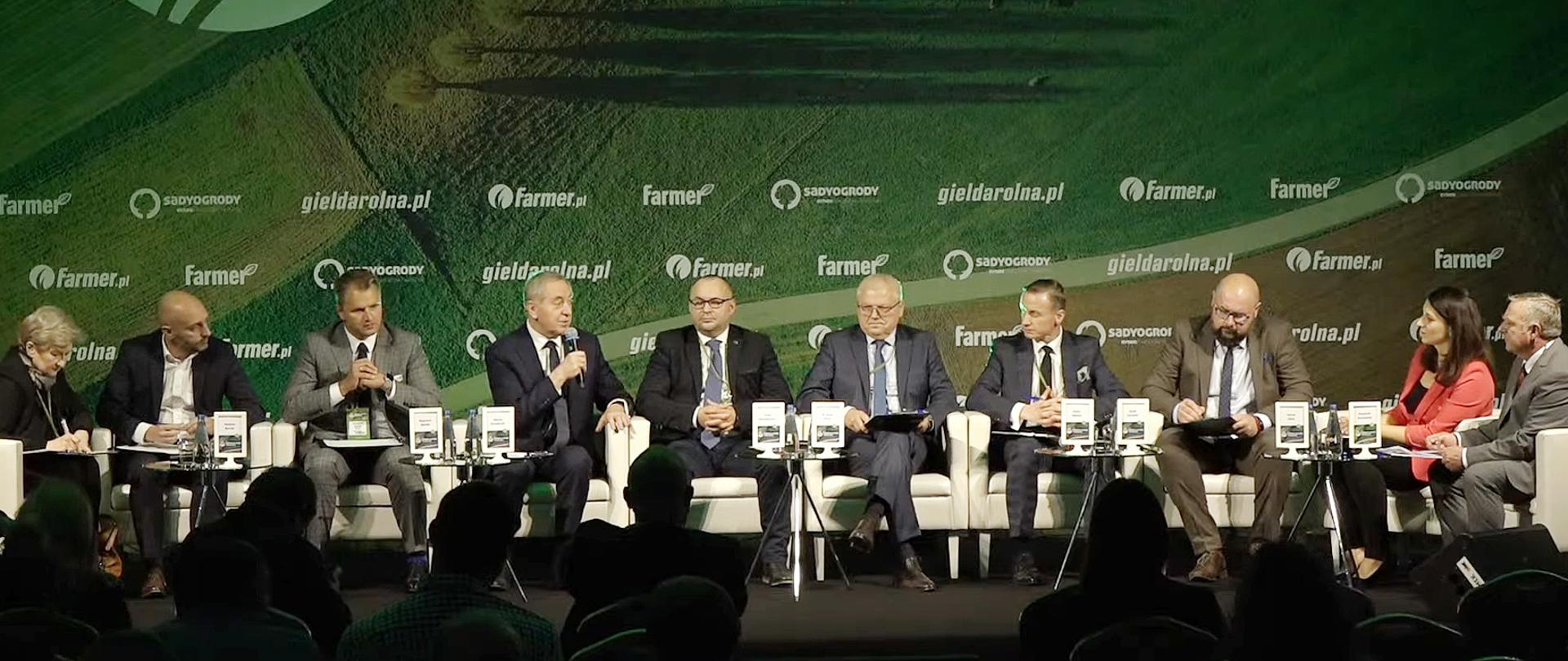 Wicepremier H. Kowalczyk z mikrofonem w prawej ręce siedzący wśród uczestników debaty.