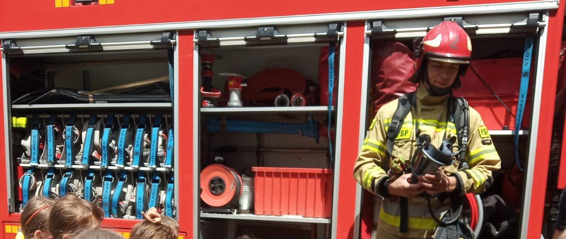 Na zdjęciu samochód ratowniczo-gaśniczy z JRG. Samochód czerwony, lampy niebieskie, ostrzegawcze. Wokół ludzie, którzy uczestniczą w pokazie sprzętu. Zdjęcie wykonane w ciągu dnia. W tle widać zielone korony drzew.