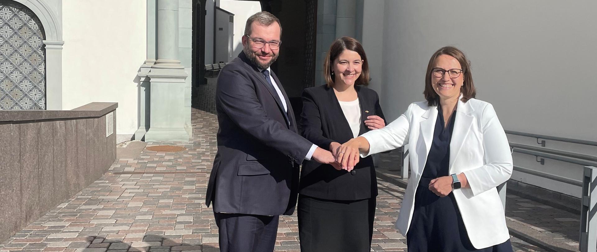 Trzy osoby stoją obok siebie i trzymają się za ręce.