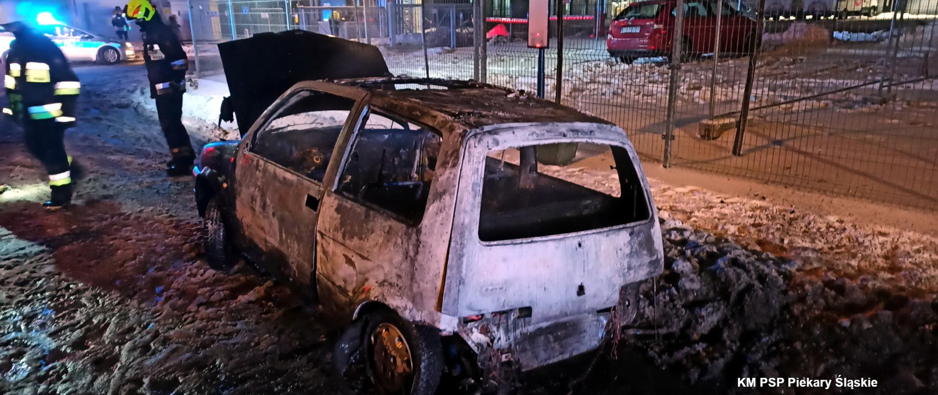 Zdjęcie przedstawia wrak spalonego samochodu marki Fiat Seicento. Obok wraku stoi dwóch strażaków w ubraniach specjalnych.