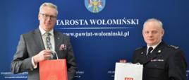 Na zdjęciu po lewej stronie Starosta Wołomiński Adam Lubiak, natomiast po prawej stronie pełniący obowiązki Komendanta Powiatowego Państwowej Straży Pożarnej w Wołominie starszy brygadier Bogusław Majchrzak. Obaj trzymają w ręku torby z materiałami promocyjnymi Starostwa i Komendy powiatowej. W tle niebieska plansza promocyjna Starostwa Powiatowego w Wołominie z herbem powiatu oraz adresem e-mail. 