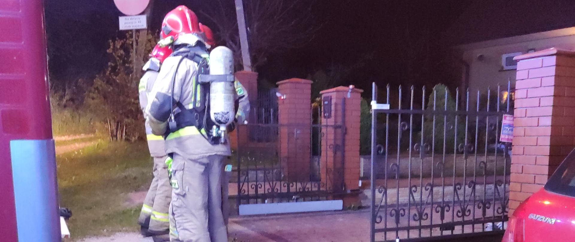 Zdjęcie przedstawia dwóch ratowników, w tym jeden zabezpieczony w aparat ochrony układu oddechowego stojących przed bramą budynku w którym wybuchł pożar. 