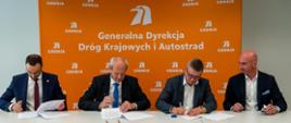 Moment podpisania jednego z 18 aneksów waloryzacyjnych. 4 elegancko ubranych mężczyzn siedzących obok siebie podpisuje dokumenty. W tle pomarańczowa ścianka z logotypami i nazwą GDDKiA.