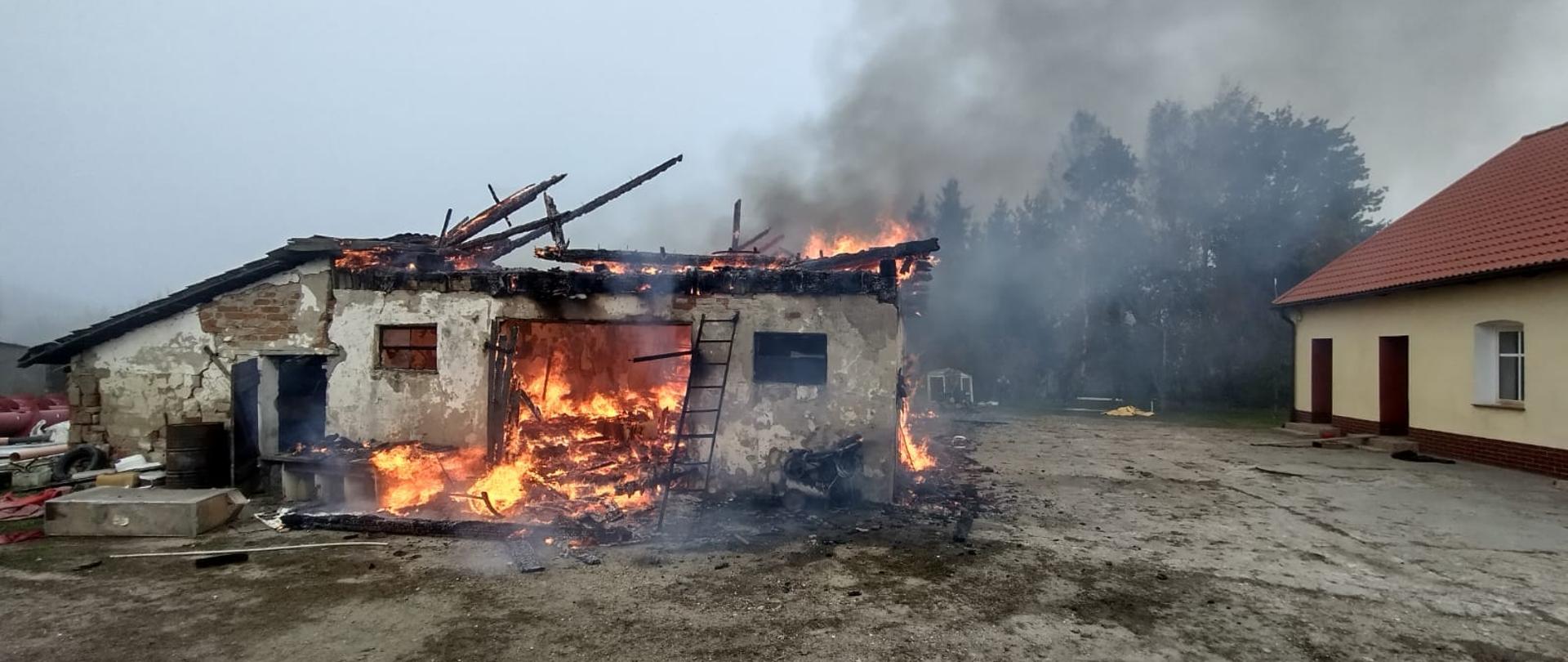 Budynek gospodarczy w płomieniach, unosi się dym. Z prawej strony budynek mieszkalny.