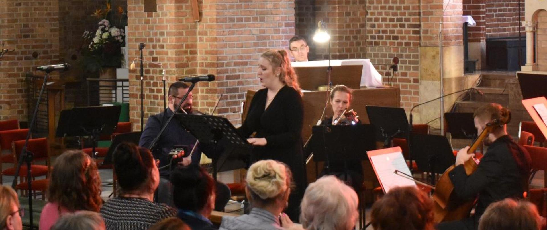 na zdjęciu, w kościele OO. Franciszkanów, podczas koncertu w ramach festiwalu Musica Classica w Jaśle widnieje solistka sopranistka i towarzyszący jej zespół kameralny (organy, dwoje skrzypce i wiolonczela) oraz zgromadzeni słuchacze. Kolorystyka zdjęcia jest biało-beżowo-brązowo-czarna. 